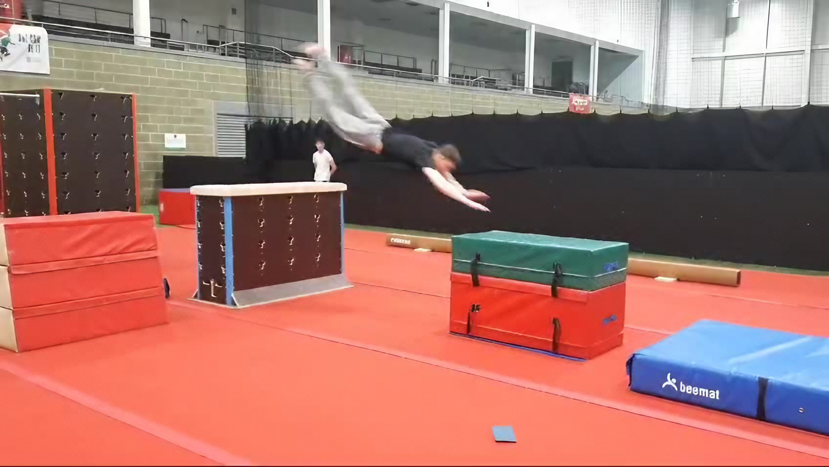 A person is performing a parkour jump in an indoor gym, leaping over padded obstacles with a red-covered floor and a wall behind.