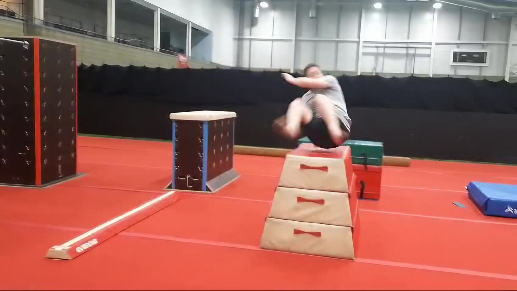 A person is mid-air while jumping over padded obstacles set up in an indoor gym with red flooring.